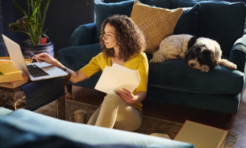 Eine Frau sitzt in ihrem Wohnzimmer und vergleicht Angebote im Internet. Hinter ihr liegt ein Hund auf der Couch.