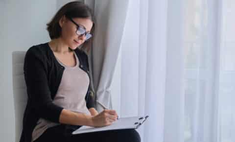 Eine Frau mit einer Brille sitzt vor einem Vorhang und macht sich auf einem Klemmbrett Notizen